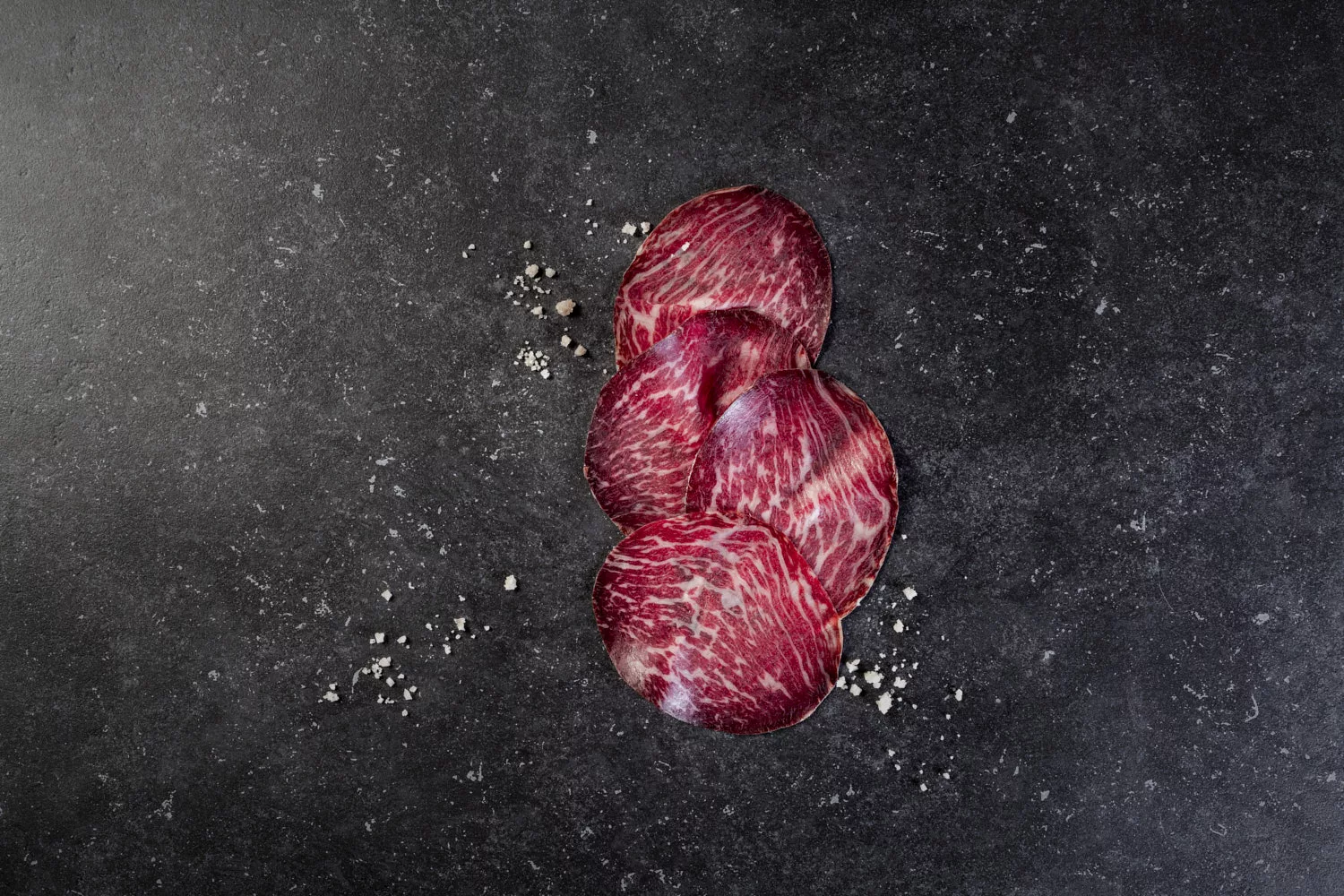 three slices of dried wagyu ham with seasalt on a black stone background