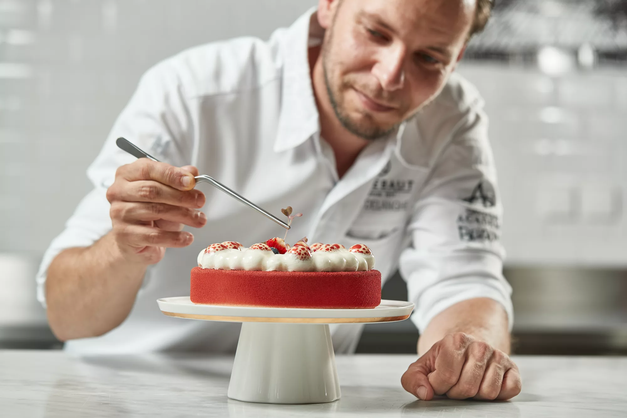 Culinary documentary and editorial chef portrait capturing the precision craftsmanship of a pastry chef fine-tuning a high-end red berry dessert on a white cake stand against a softly lit professional kitchen background.