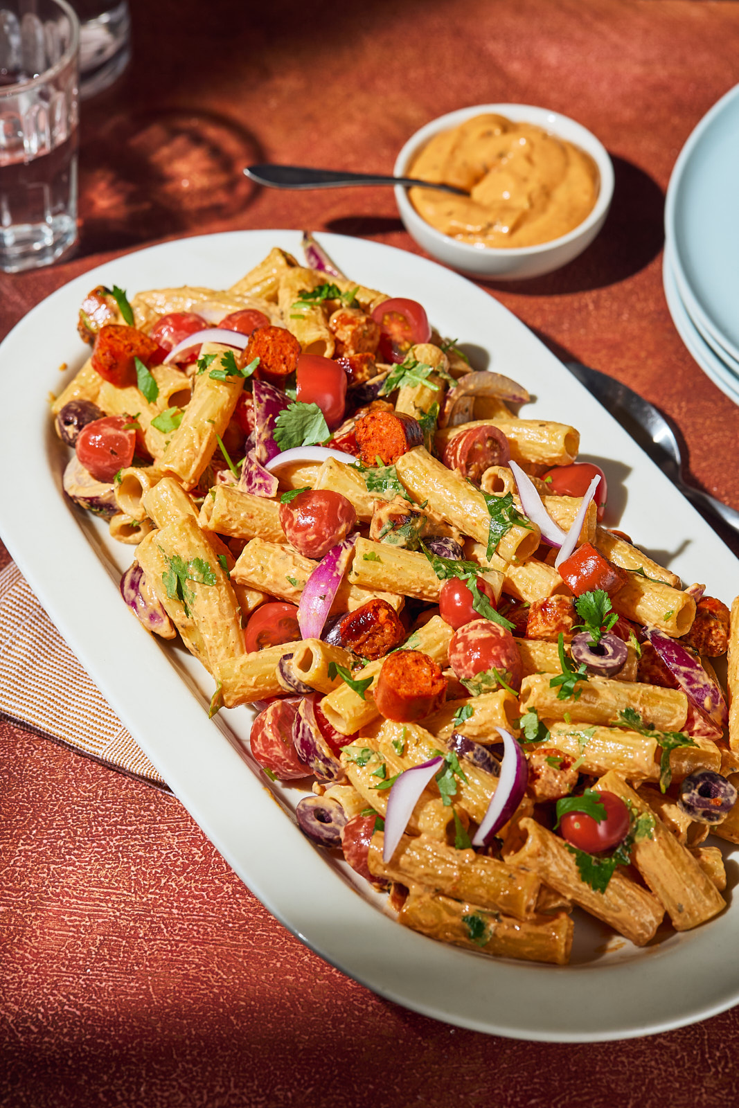 High-angle shot of a white serving platter piled high with creamy rigatoni pasta salad featuring chorizo, cherry tomatoes, sliced red onion, olives, and fresh herbs, alongside a small dish of orange dip.