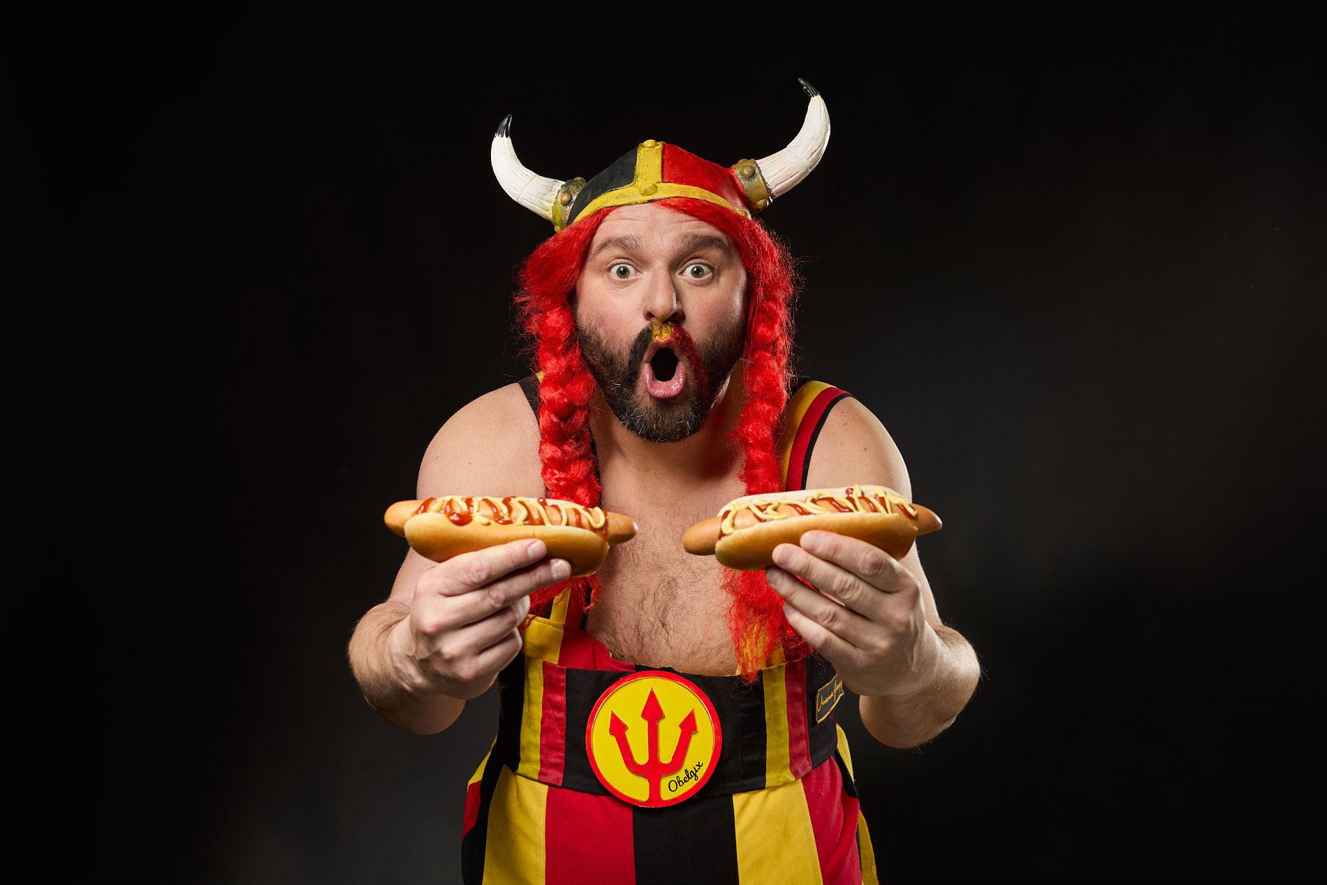 Studio portrait of Belgian supporter Obelgix with an open-mouthed expression, dressed in red-and-yellow striped overalls and a horned helmet with red braided pigtails, presenting a pair of hot dogs topped with condiments toward the camera.