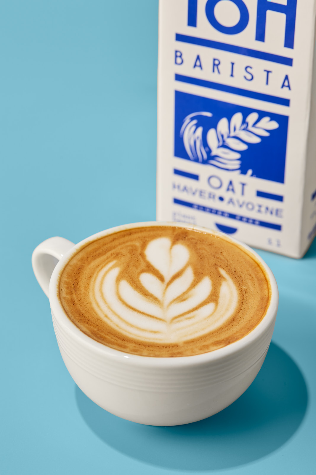 Close-up shot of a white cup filled with a cappuccino featuring a tulip latte art design, placed beside a white and blue TipToh Barista Oat milk carton on a solid bright blue surface.